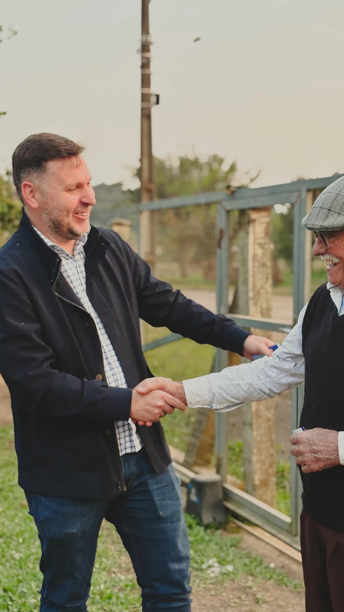 Gabriel Diedrich aperta a mão de um senhor de boina e óculos em frente a um portão, numa propriedade rural, no fim da tarde.